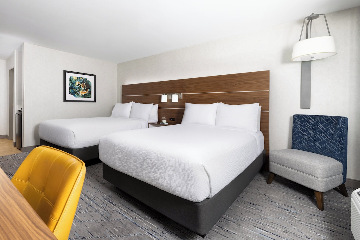 Quarto de hotel com uma cama grande, uma cama menor, uma escrivaninha, uma cadeira e um abajur.