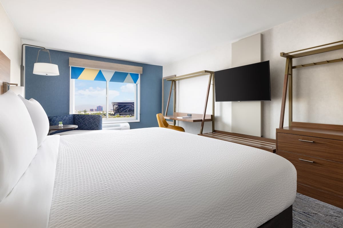 A hotel room with a large bed, a desk, a chair, a TV, and a window with a view of a cityscape.