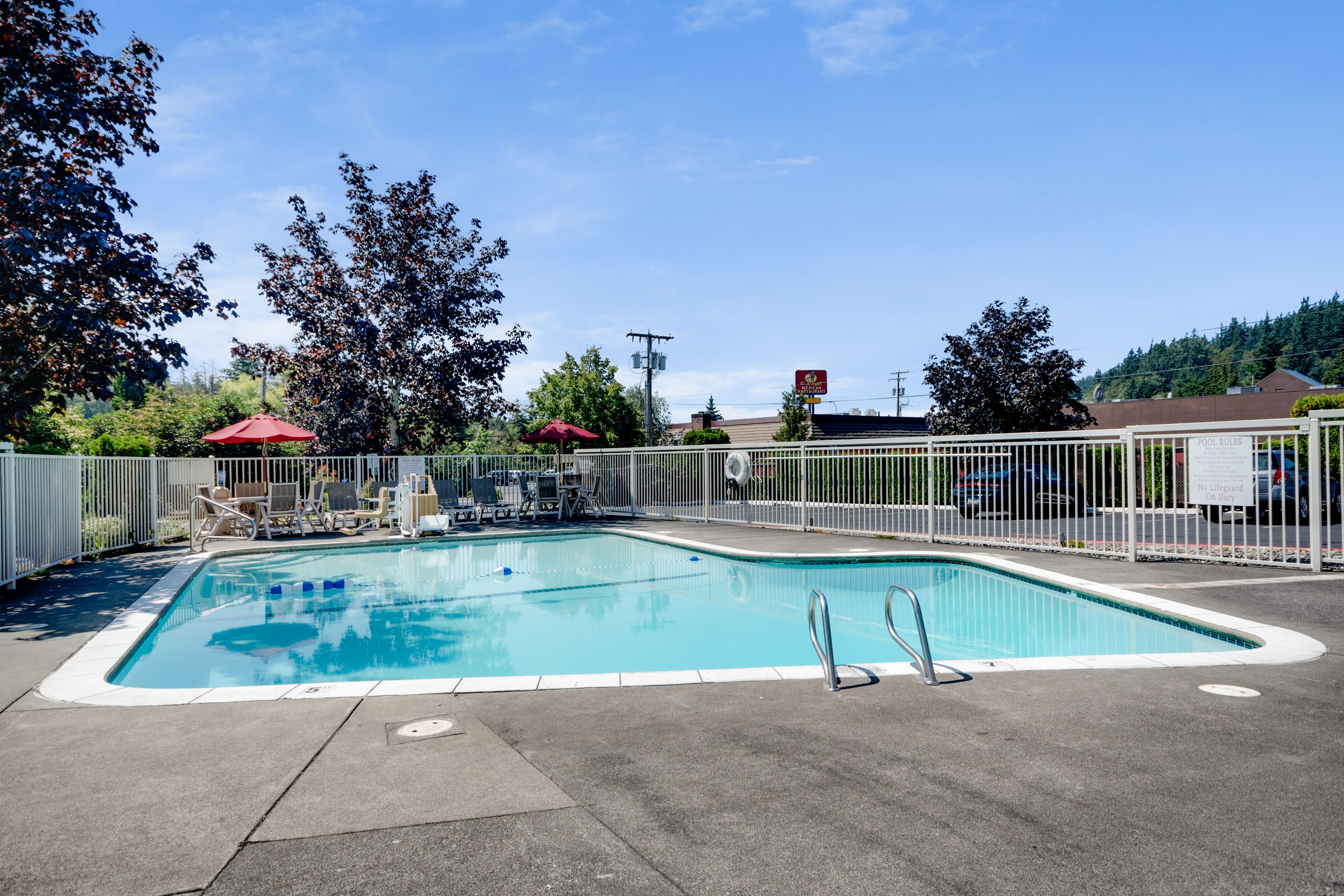 A rectangular outdoor swimming pool with a white fence, surrounded by trees and a clear sky.
