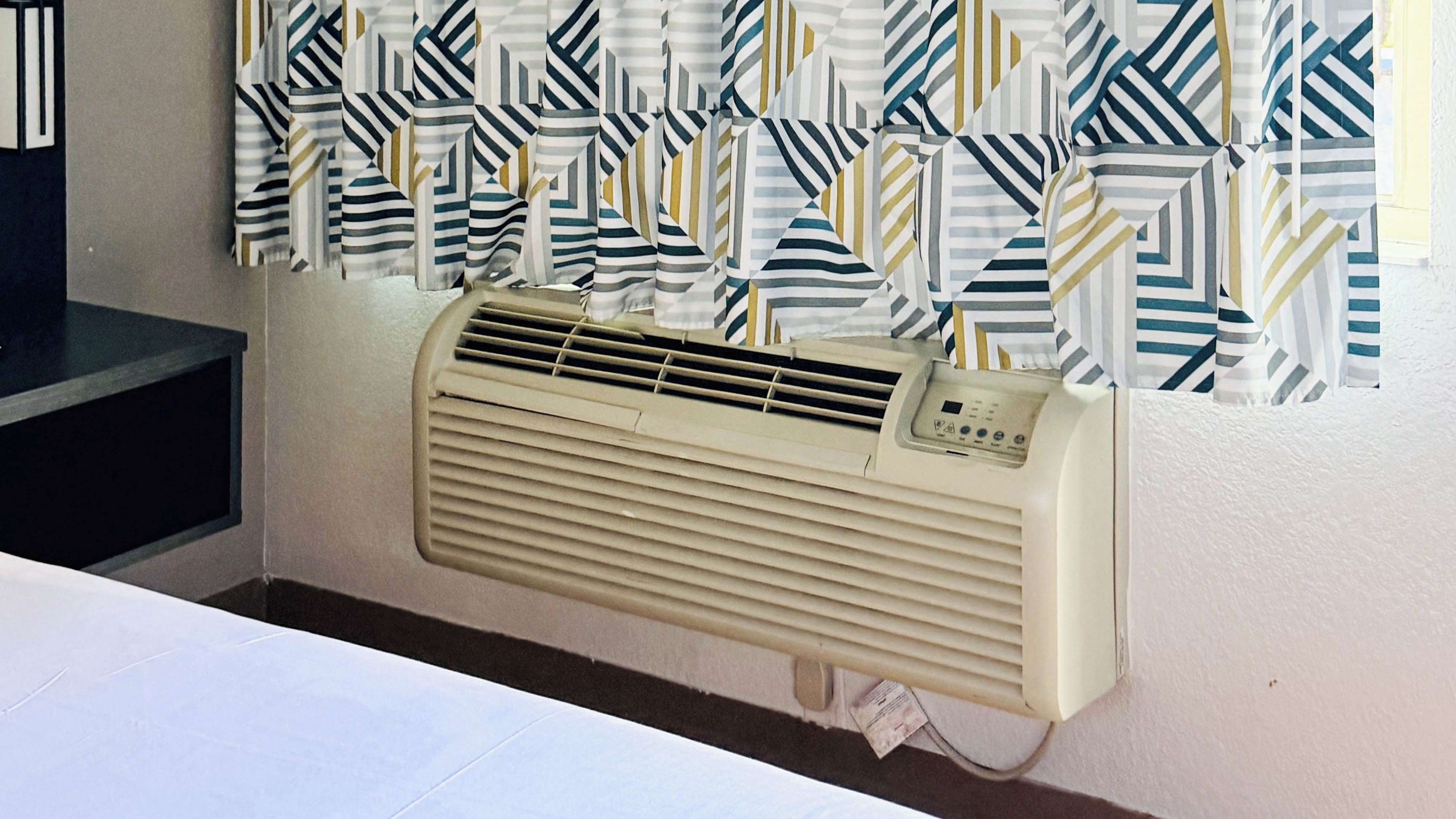 A room with a beige air conditioning unit mounted on the wall, a window with striped curtains, and a dark-colored table.