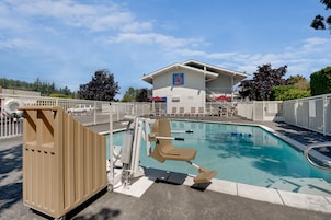 A swimming pool with a lifeguard chair and a wooden structure, surrounded by a fence, with a building in the background.