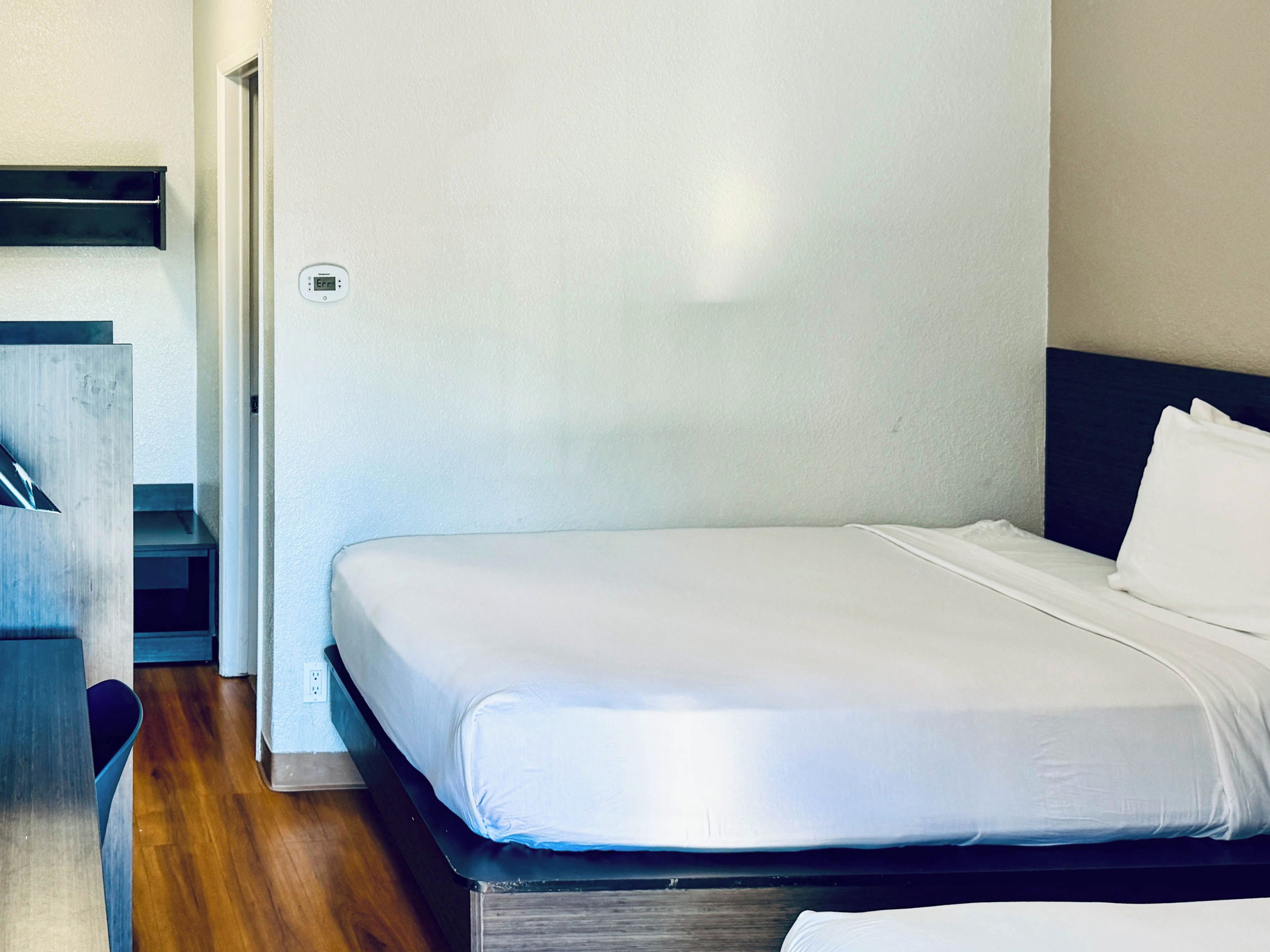 Een eenpersoonsbed met een witte matras en kussen, een houten vloer en een witte muur met een stopcontact.