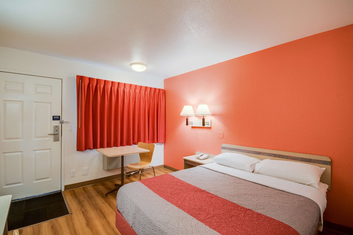 Habitación de hotel con cama, escritorio, silla, mesita de noche y lámpara de pared.