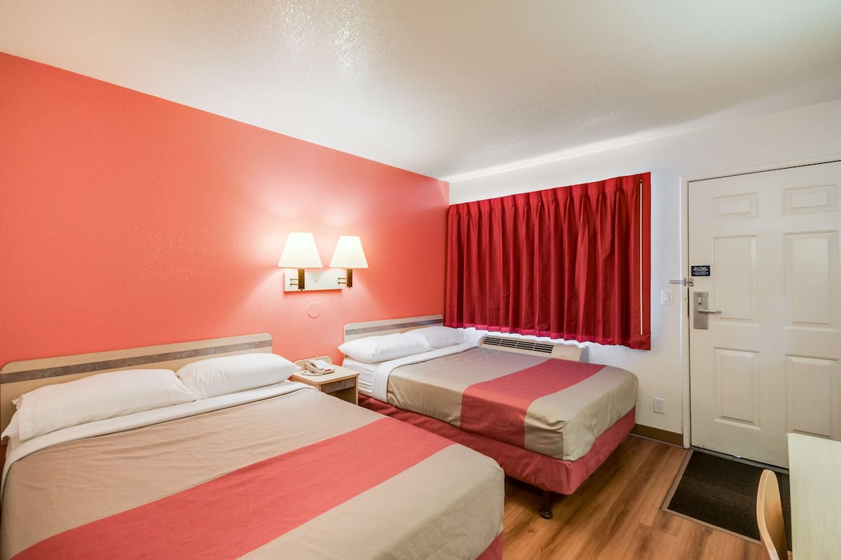 Habitación de hotel con dos camas, una pared roja, una puerta blanca y una mesita de noche con lámpara.