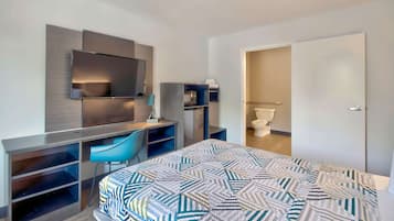 A modern hotel room with a bed, a desk with a chair, a TV mounted on the wall, and a bathroom visible through an open door.