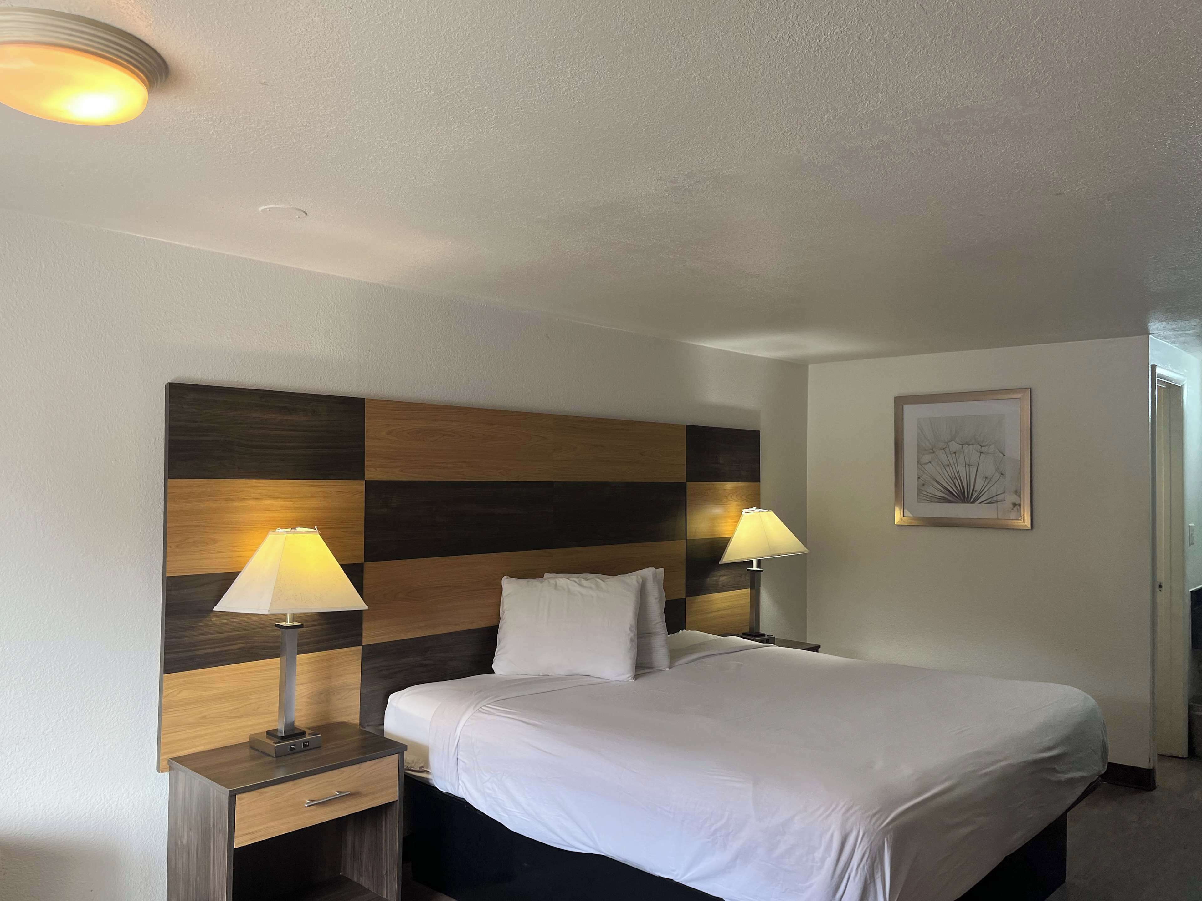 A hotel room with a large bed, two bedside tables with lamps, a headboard with a wood panel design, and a framed artwork on the wall.