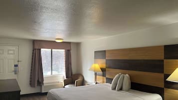 Et hotelværelse med seng, stol, skrivebord, lampe og et vindue med gardiner.