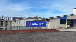 A motel with a 'NOW OPEN' sign, a fenced parking area, and a clear blue sky. - Motel 6 Cross Lanes, WV - Goff Mountain Rd (Cross Lanes)