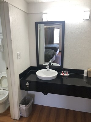 Un baño de hotel con un lavamanos blanco, encimera negra y un espejo. Se ve un inodoro a la izquierda y un cubo de basura debajo del lavamanos.