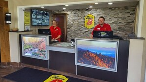 A reception desk with two employees, a monitor displaying a scenic image, and a logo on the floor mat.
