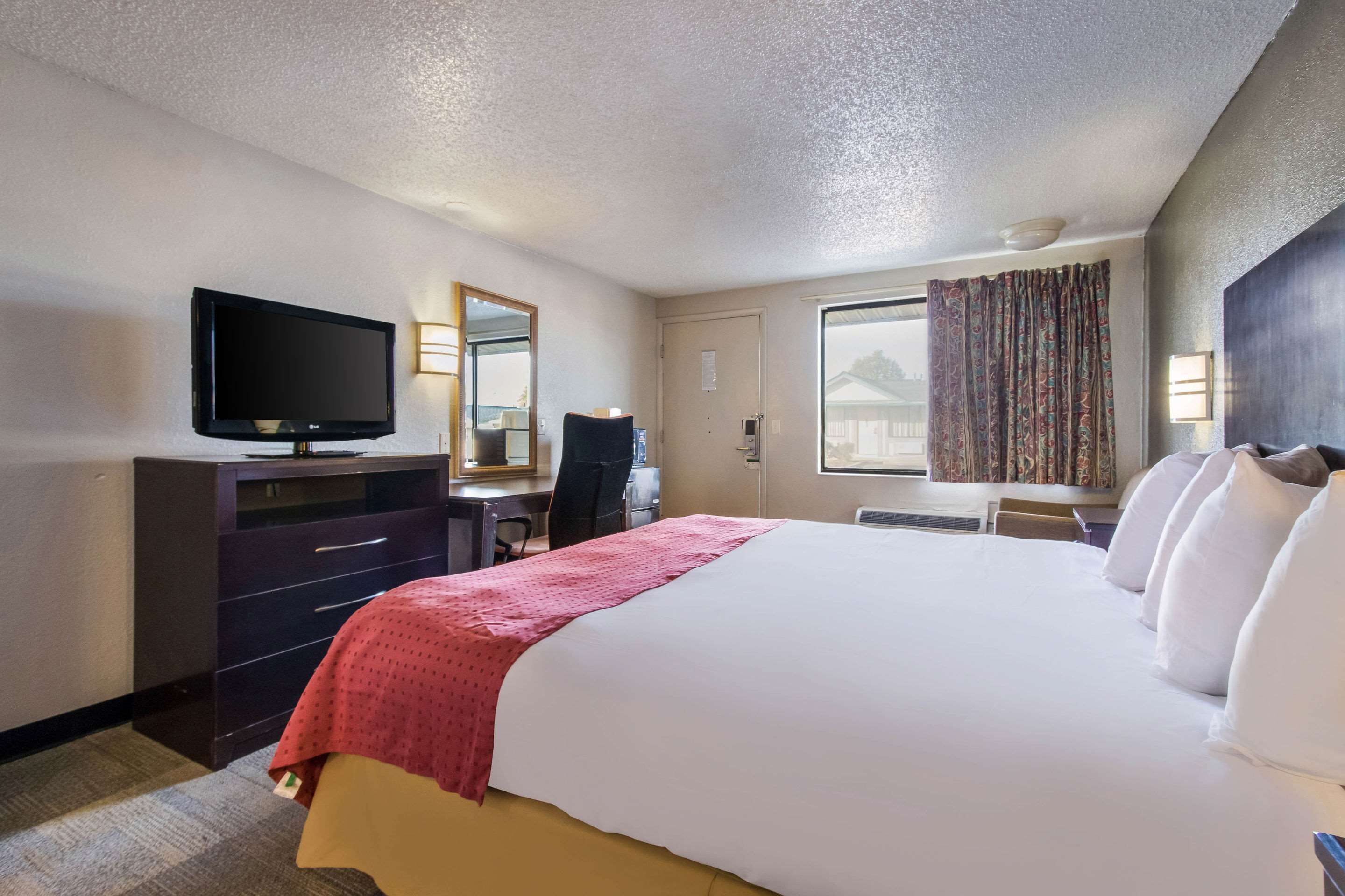 A hotel room with a large bed, a desk with a chair, a television, a window with curtains, and a door.