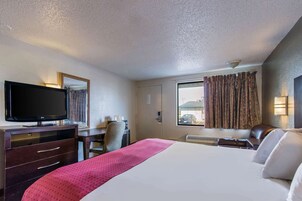 Habitación de hotel con cama, escritorio y silla, televisión, ventana con cortinas y puerta.