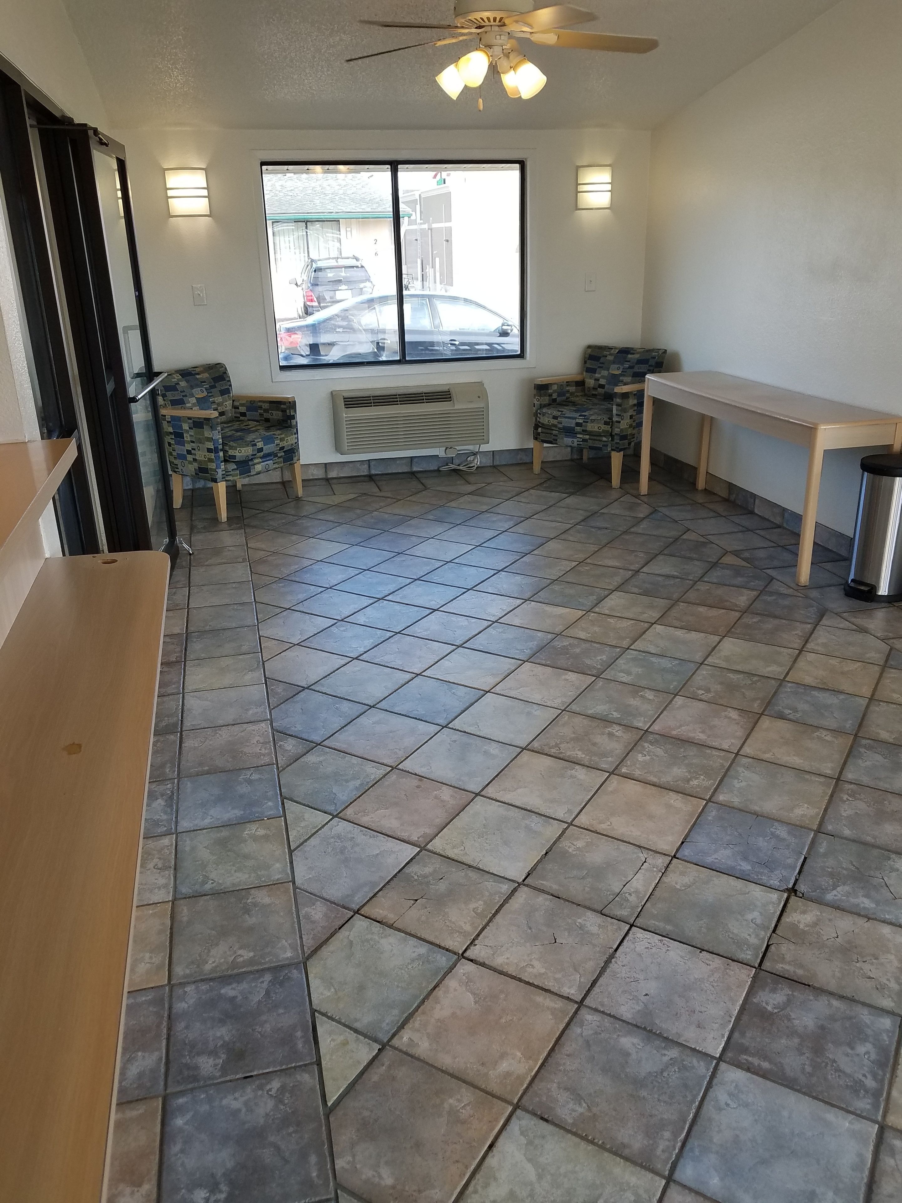 A room with a tiled floor, a ceiling fan, two patterned chairs, a wooden table, and a window with a view of parked cars outside.