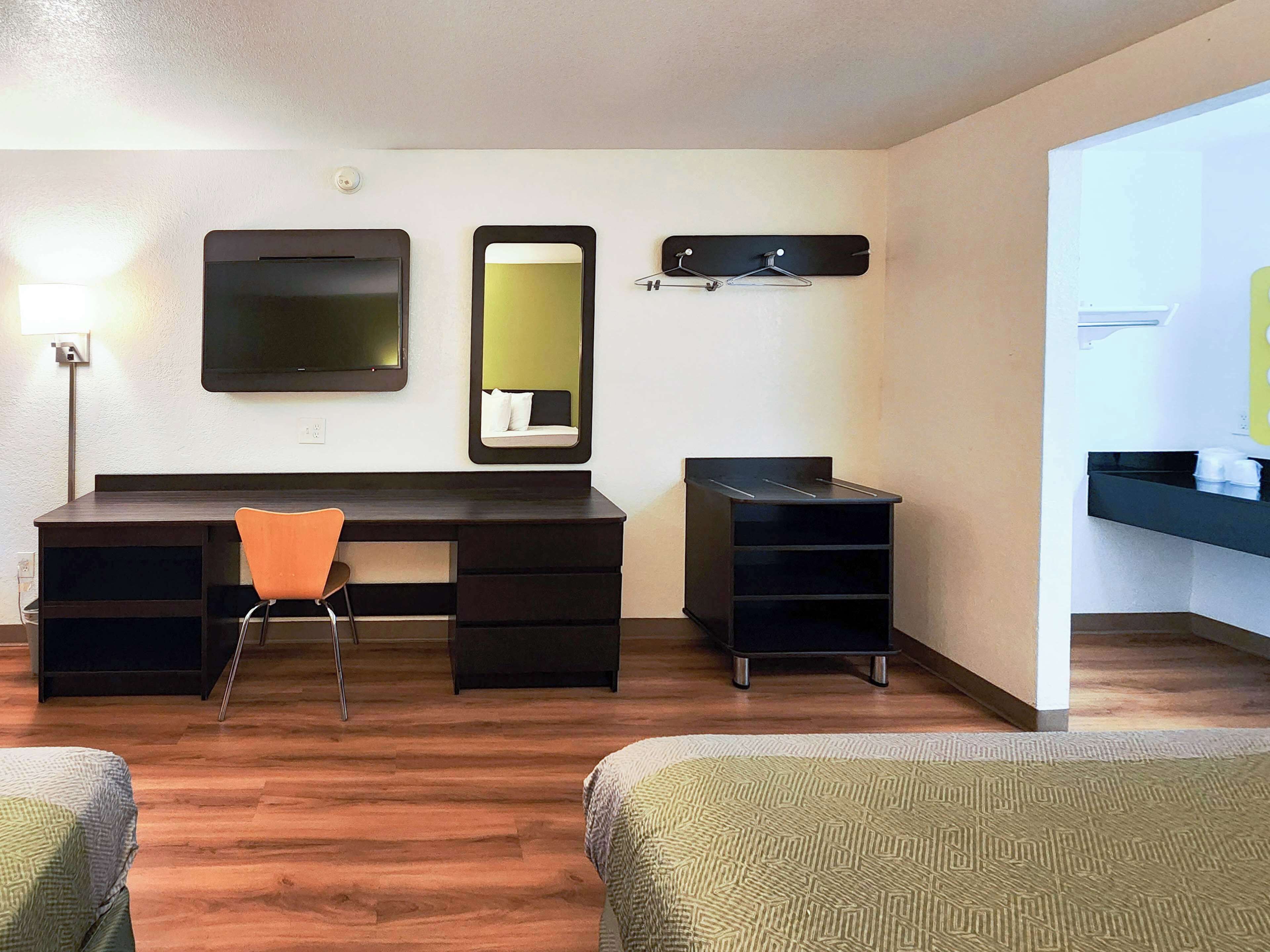 A hotel room with a desk, two chairs, a mirror, a shelf with cups, and a bed.