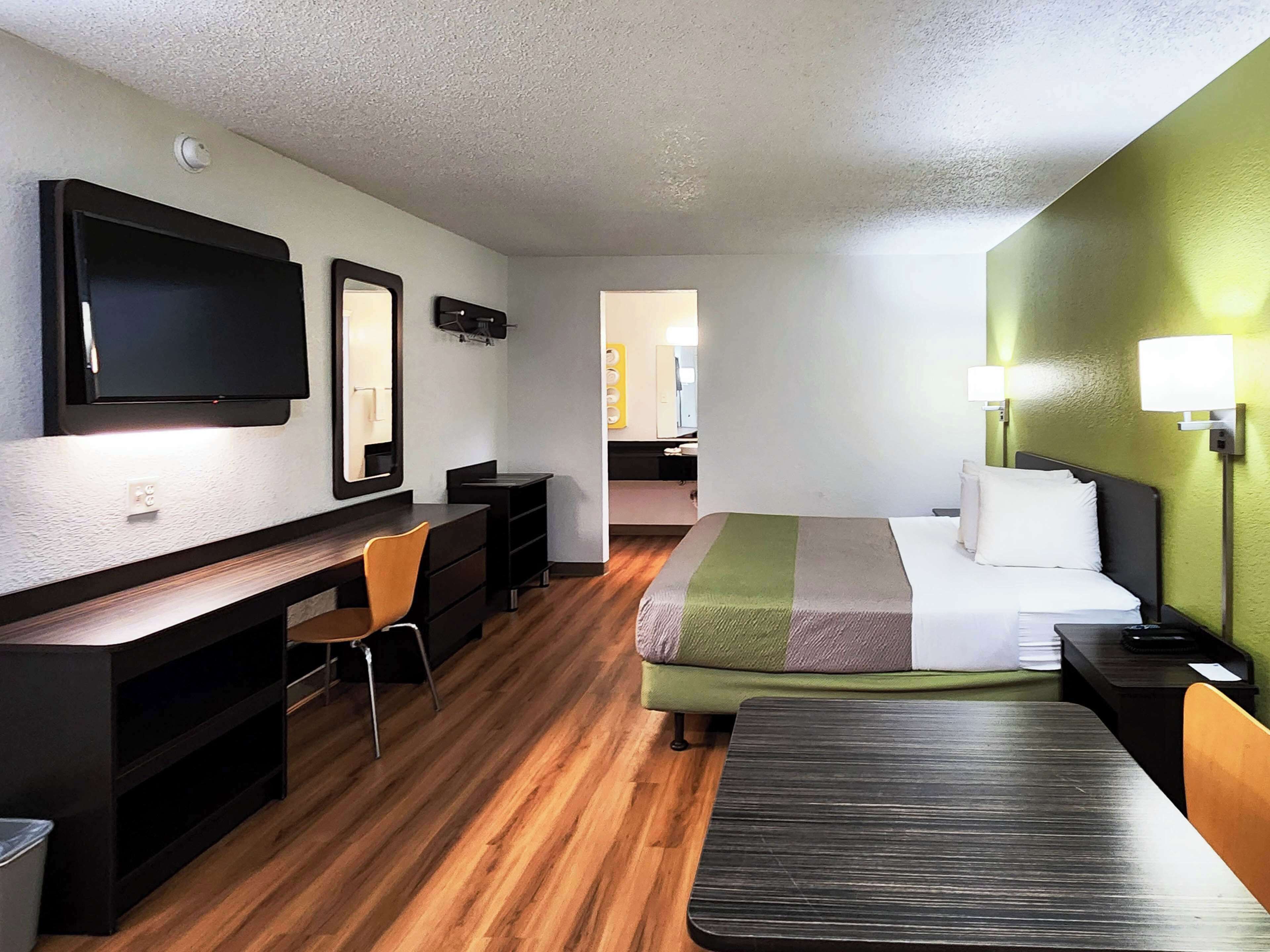Une chambre d’hôtel avec un plancher en bois, un bureau avec une chaise, un lit recouvert d’une couverture à rayures vertes et blanches, et un téléviseur fixé au mur.