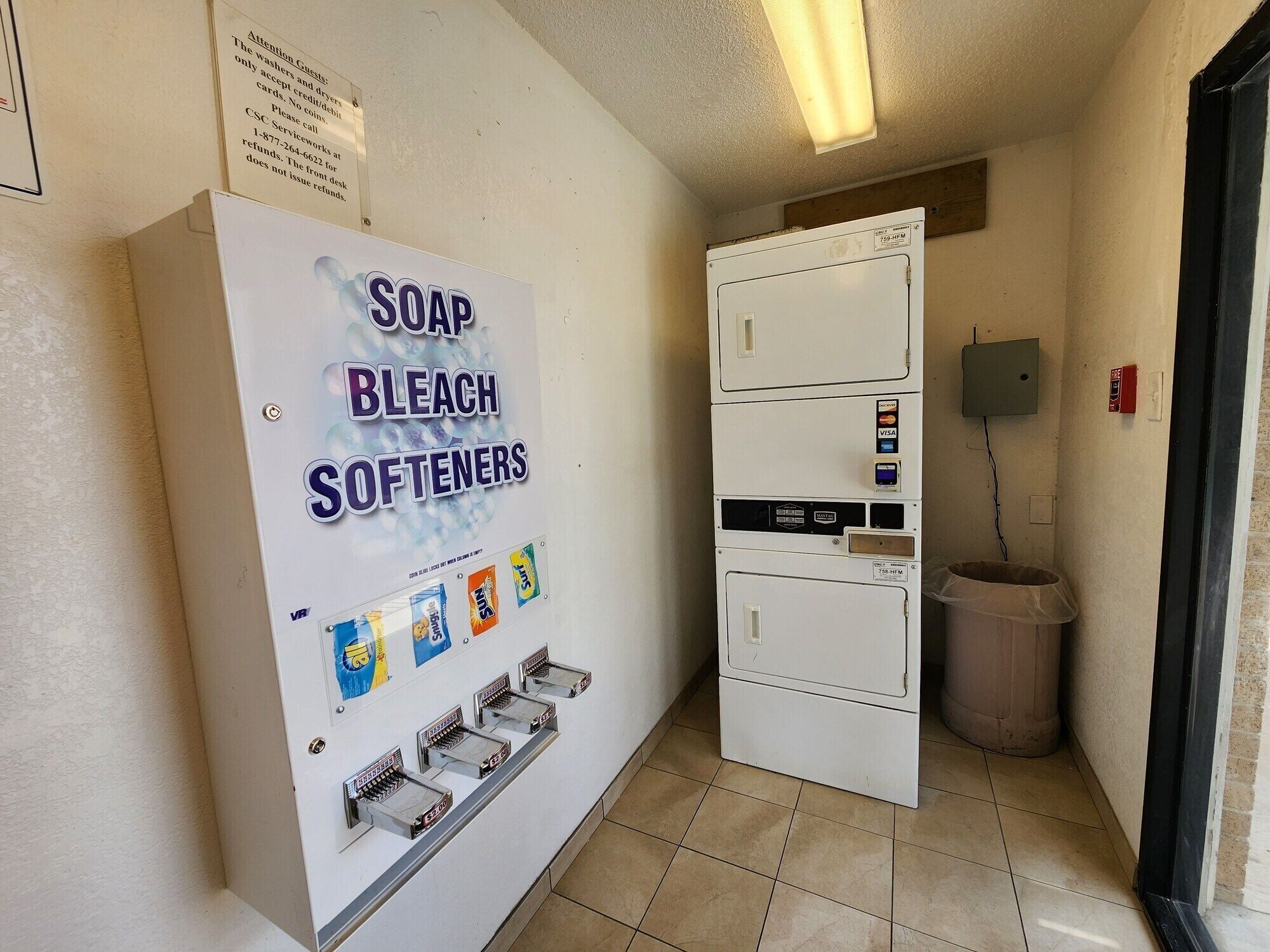 A utility room with a soap, bleach, and softener dispenser, a small trash bin, and a fire alarm box on the wall.