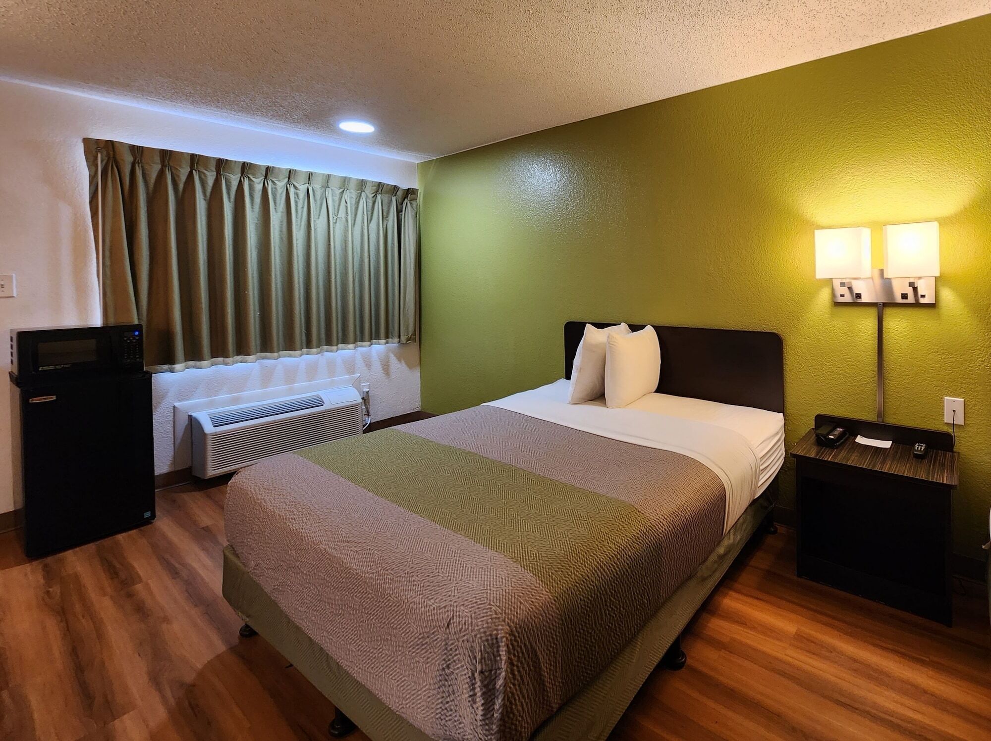 A hotel room with a bed, a nightstand, a desk with a computer, an air conditioner, and a window with curtains.