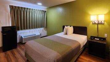 A hotel room with a bed, a nightstand, a desk with a computer, an air conditioner, and a window with curtains.