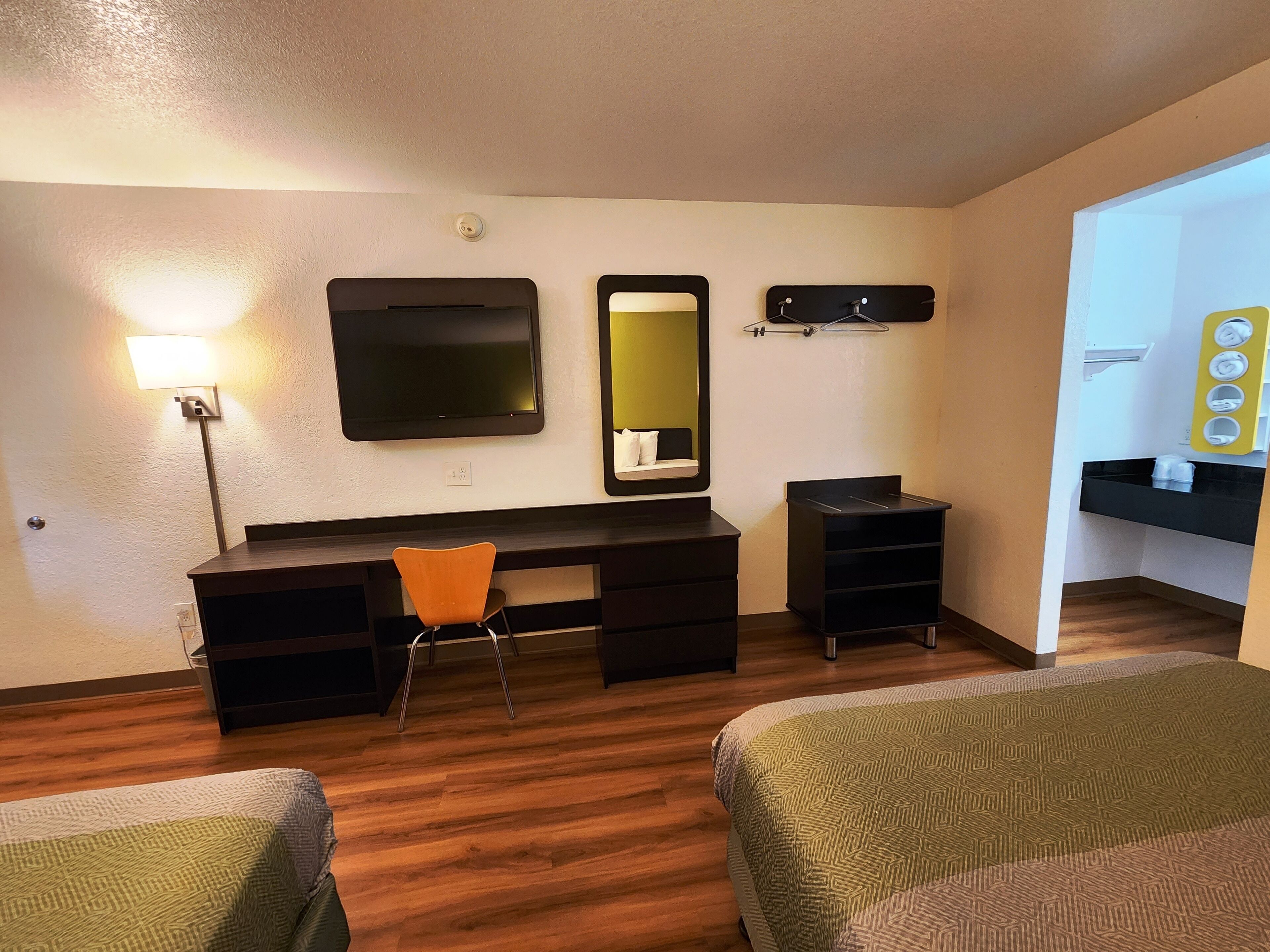 A hotel room with a desk, two chairs, a bed, a mirror, a lamp, and a small bathroom area.