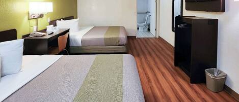 A hotel room with a large bed, a smaller bed, a desk with a lamp, a TV, and a trash can.