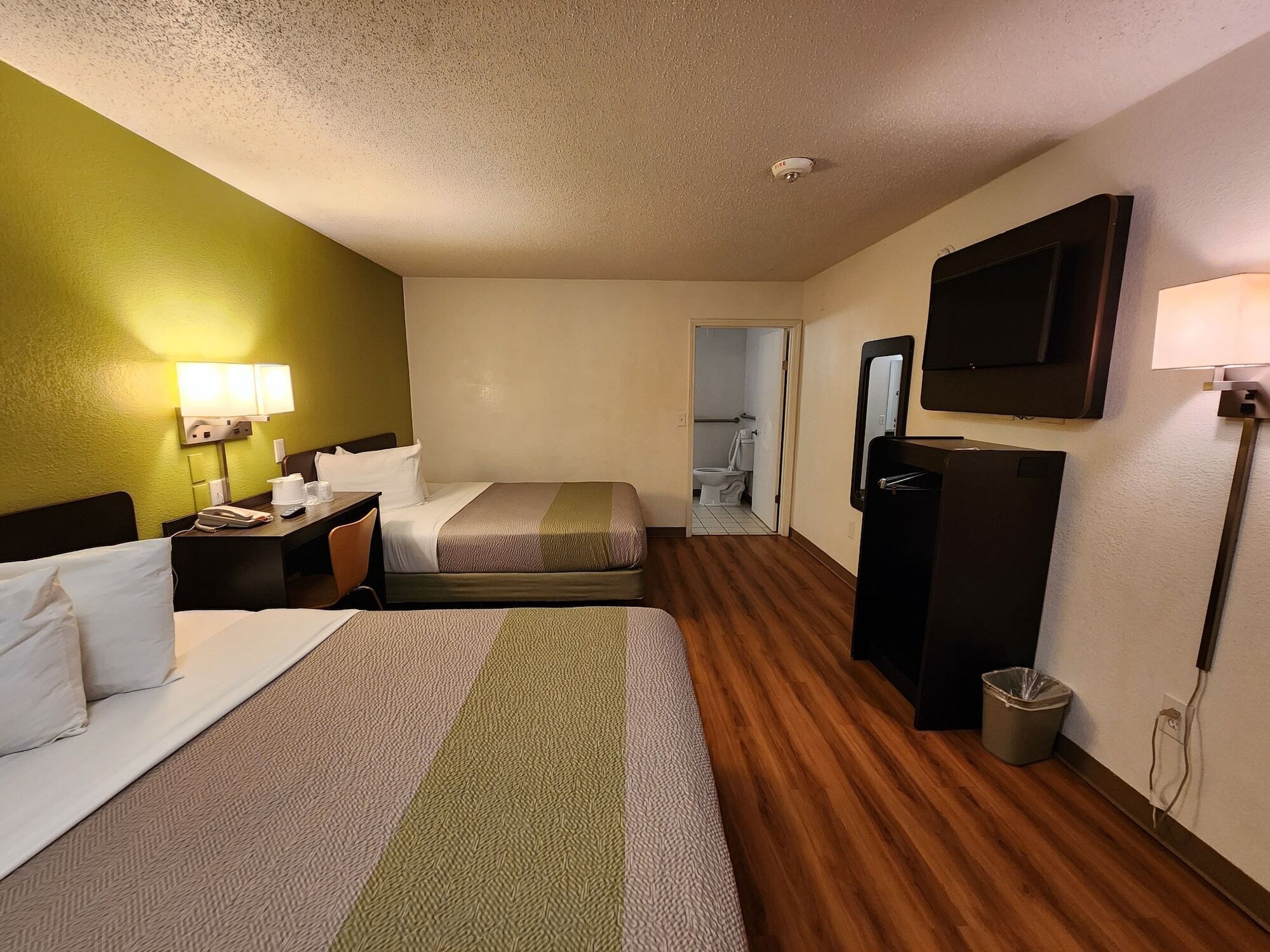A hotel room with two beds, a desk with a lamp, a TV, and a bathroom visible through an open door.