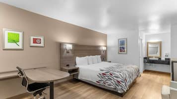 A hotel room with a bed, bedside table, desk, chair, and a bathroom with a sink and mirror.
