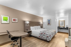 A hotel room with a bed, bedside table, desk, chair, and a bathroom with a sink and mirror. - Red Roof Inn Houston – Jersey Village (Houston)