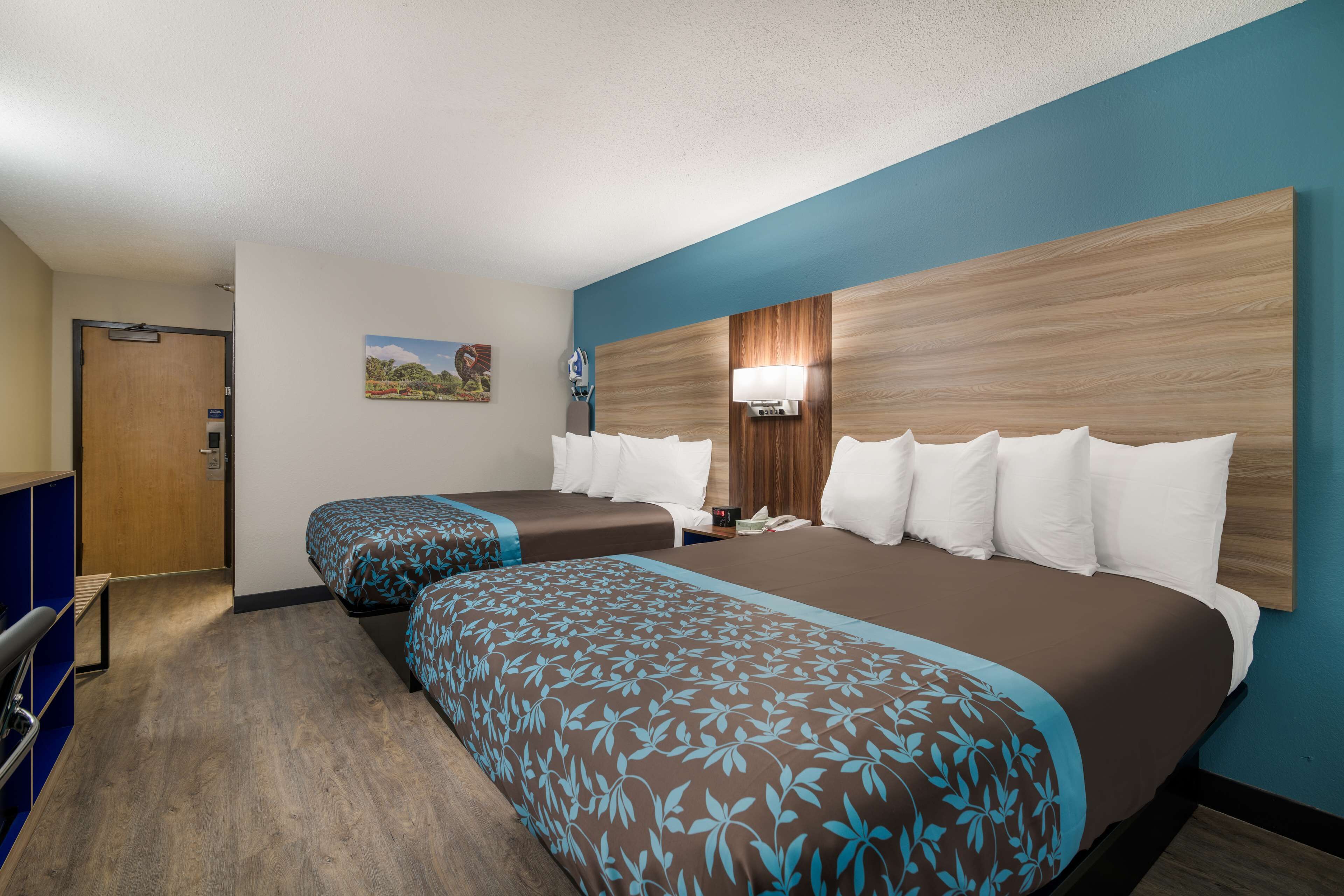 A hotel room with two beds, a wooden headboard, a nightstand, a lamp, and a small painting on the wall.