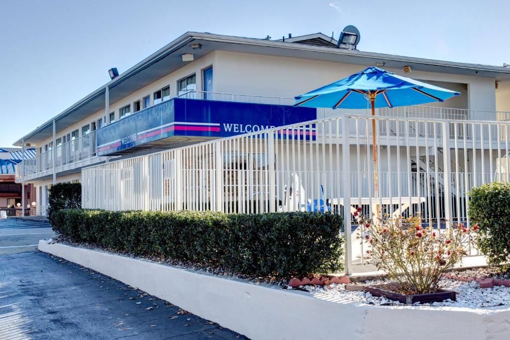 Entrada de un hotel con un letrero que dice 'WELCOME', un espacio exterior cubierto con una sombrilla azul y una valla blanca.