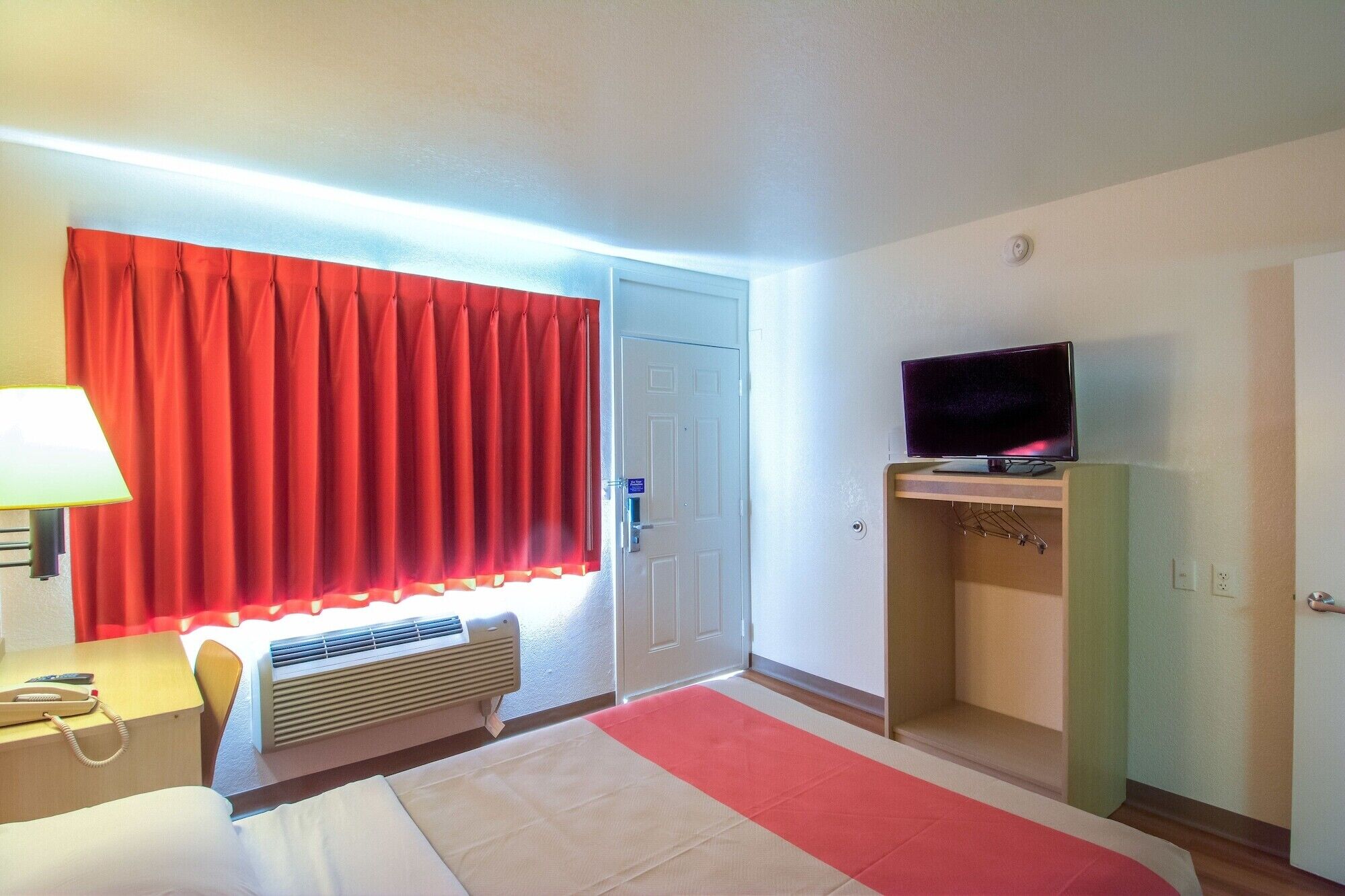 Una habitación de hotel con una cama, un escritorio con lámpara, un televisor sobre un mueble y una cortina roja.