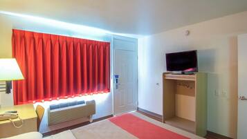 Una habitación de hotel con una cama, un escritorio con lámpara, un televisor sobre un mueble y una cortina roja.