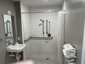 A bathroom with a white sink, a mirror, a shower with grab bars, and a towel rack. - Motel 6 Arcata, CA - Cal Poly Humboldt (Arcata)