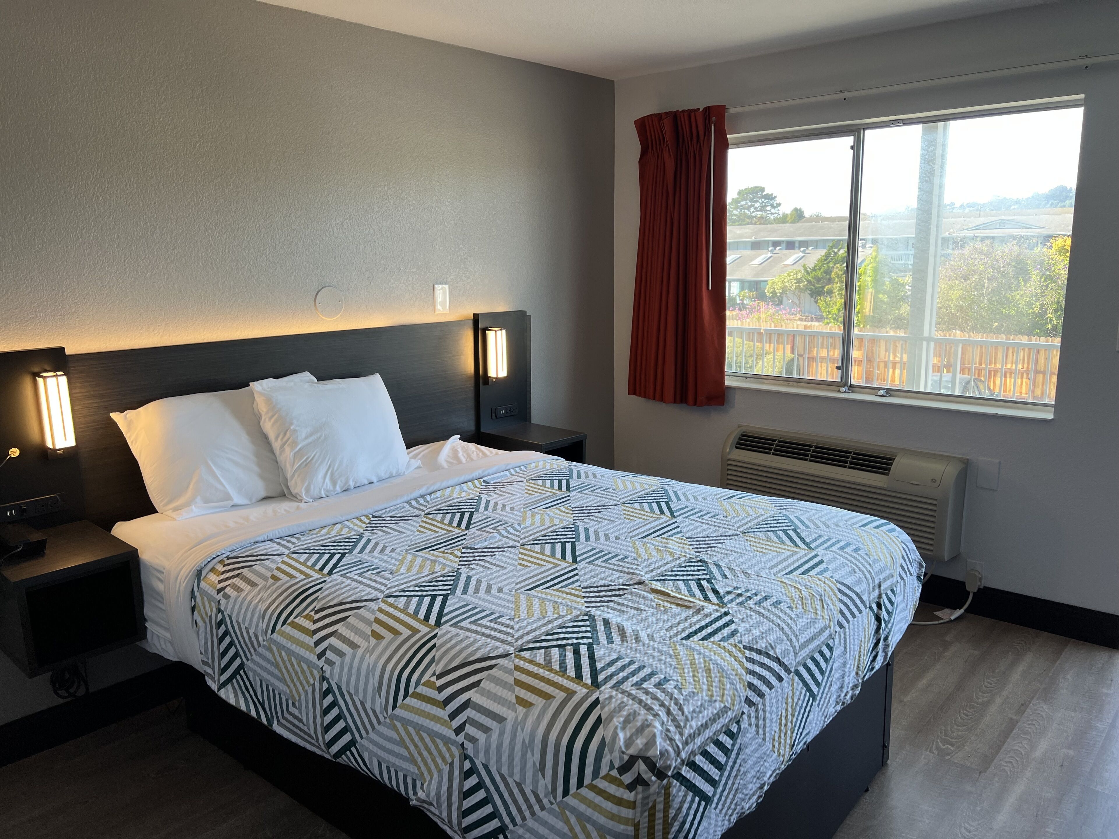 A hotel room with a large bed, two bedside tables, a nightstand, a window with a view, and a red curtain.