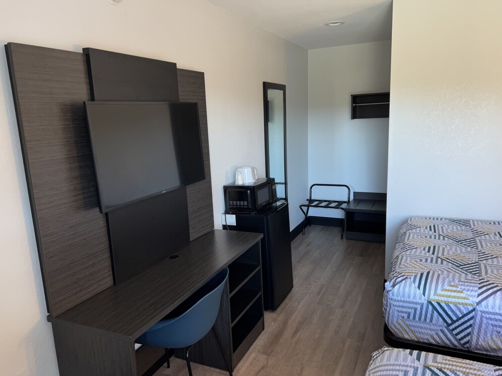 A compact hotel room with a dark wood desk, a blue chair, a bed with patterned bedding, a microwave, and a mirror.