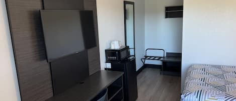 A compact hotel room with a dark wood desk, a blue chair, a bed with patterned bedding, a microwave, and a mirror.