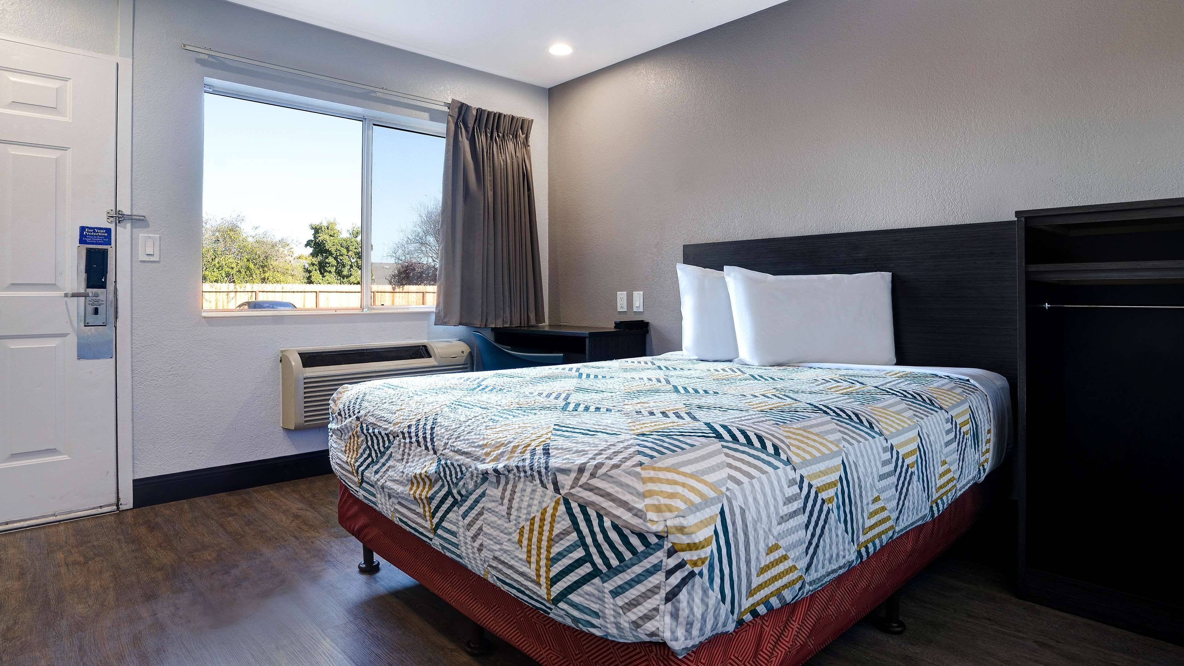 Una habitación de hotel con cama, escritorio, silla, ventana con cortinas y puerta.