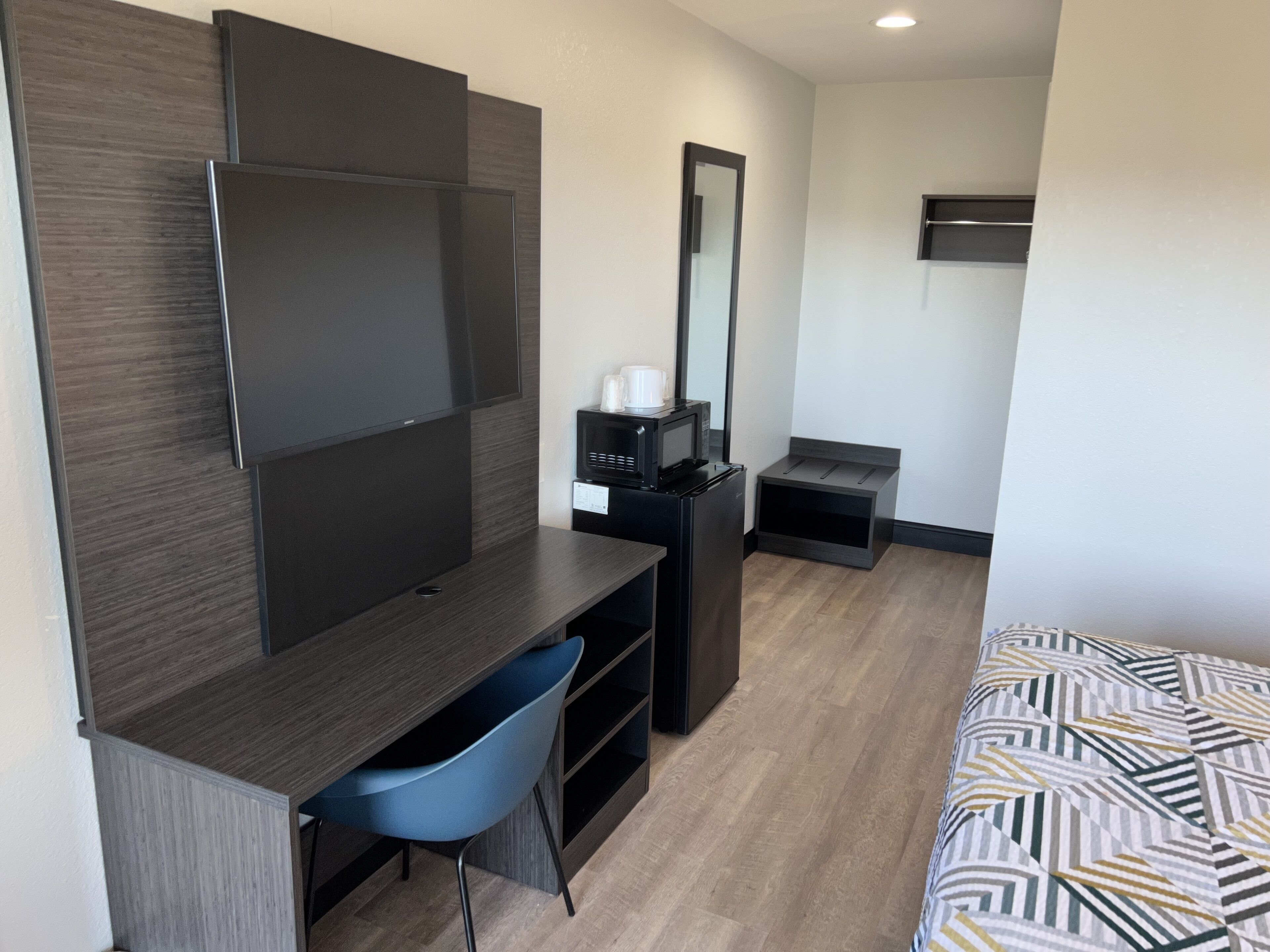A modern hotel room with a dark wood desk, a blue chair, a bed with patterned bedding, a black TV stand, and a small black cabinet.