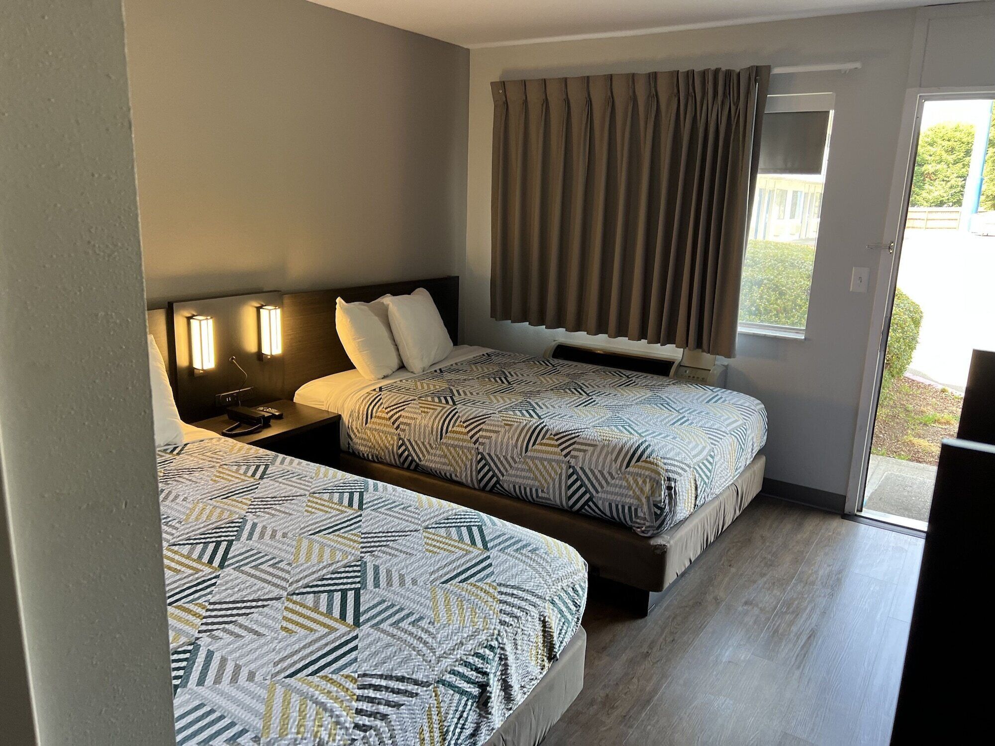 Hotellihuone, jossa on kaksi sänkyä, yöpöytä, lamppu, ikkuna verhoilla ja näkymä ulos.