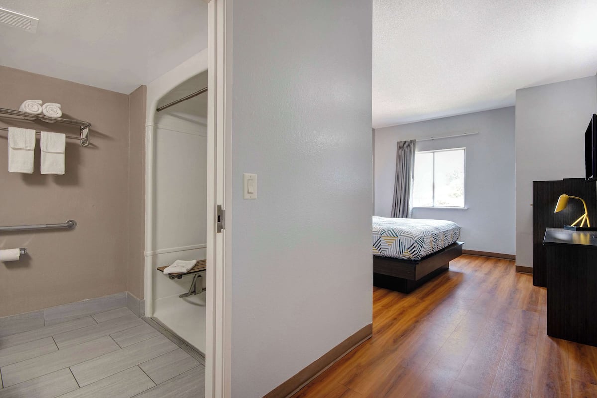 Habitación de hotel con piso de madera, cama con ropa de cama estampada, mesita de noche con lámpara y ventana con cortinas.