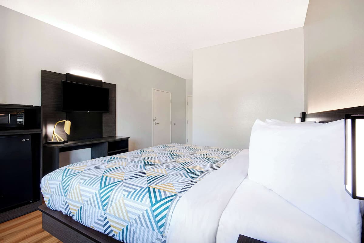 Habitación de hotel con cama, televisor montado en la pared, escritorio con lámpara y minibar.