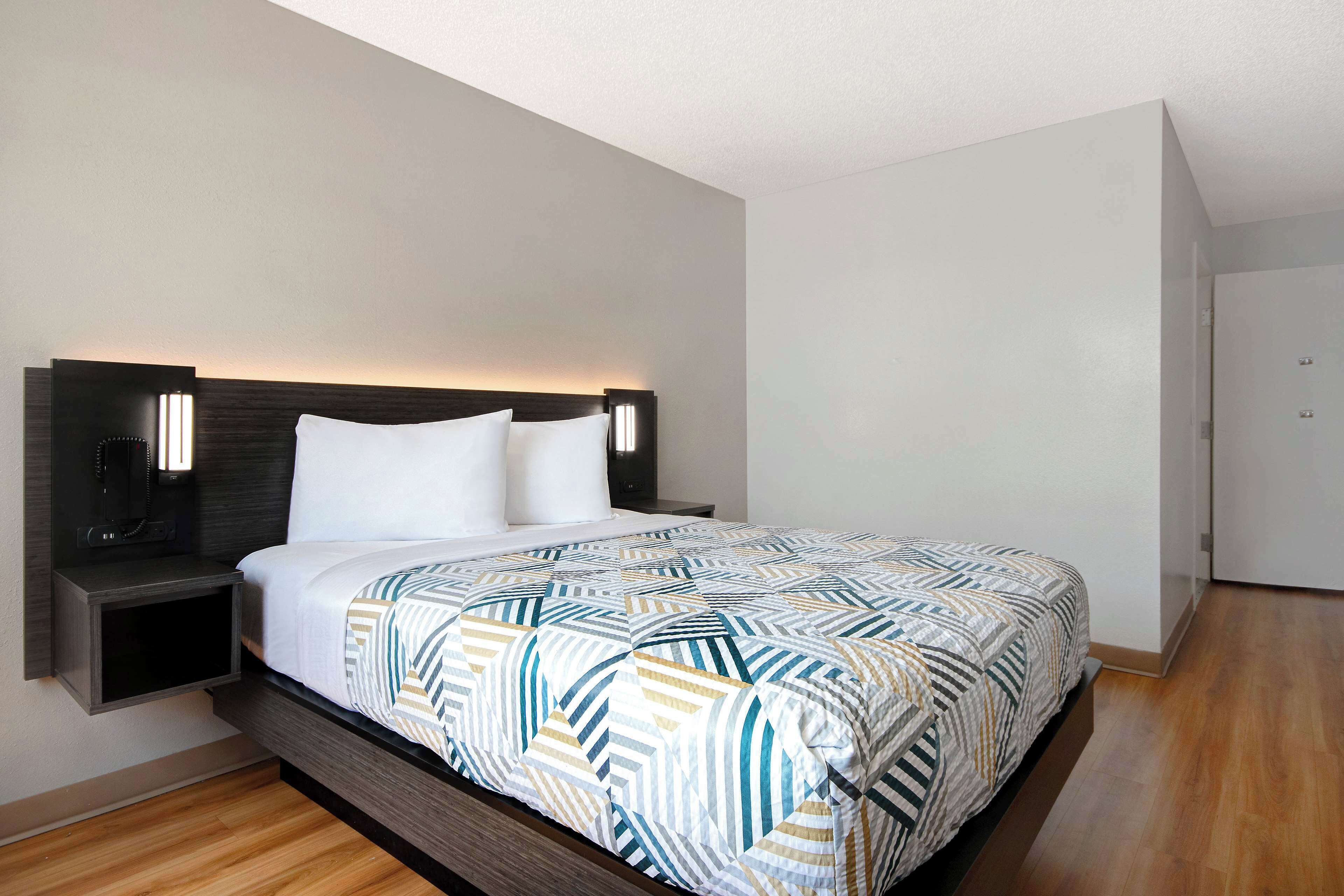 A modern hotel room with a large bed, a nightstand, and a wall-mounted lamp.
