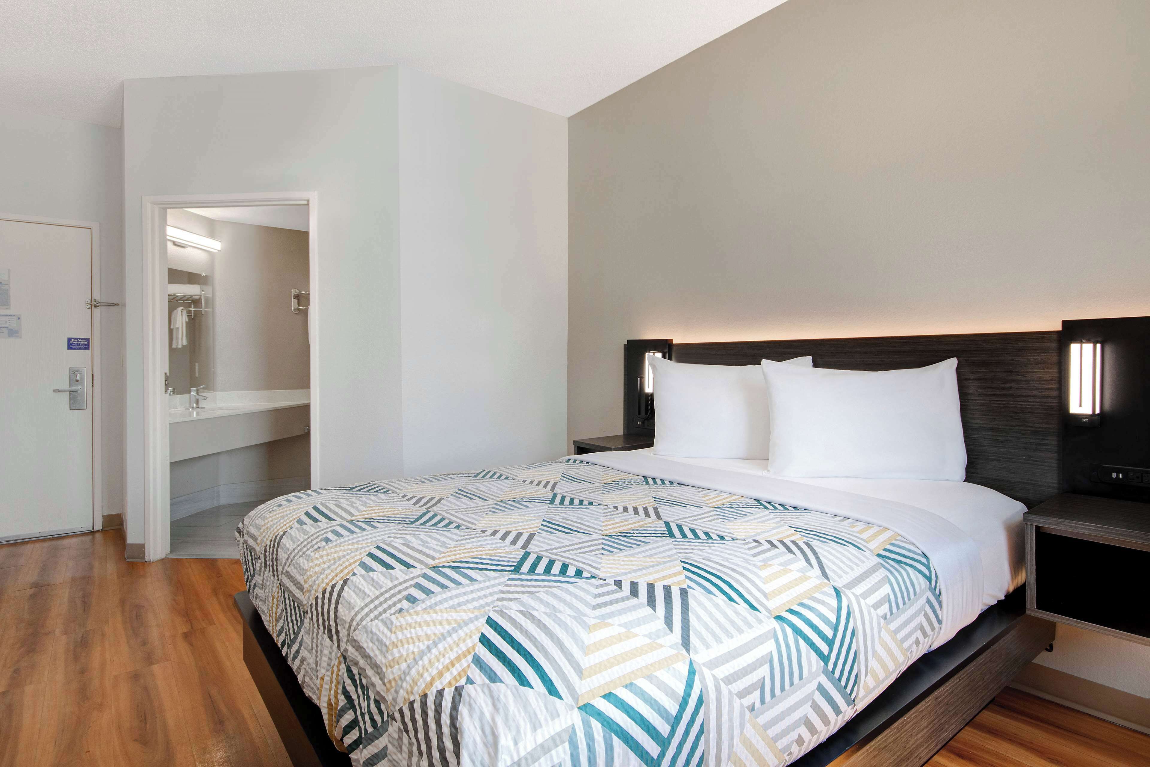 Une chambre d’hôtel avec un lit, une table de chevet, un plancher en bois et une salle de bain visible à travers une porte ouverte.
