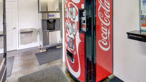 A Coca-Cola vending machine in a commercial space with a stainless steel refrigerator and a door in the background.