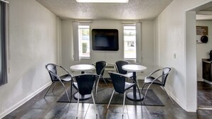 A room with a TV mounted on the wall, two windows, a seating area with four chairs and two tables, and a dark wooden floor.