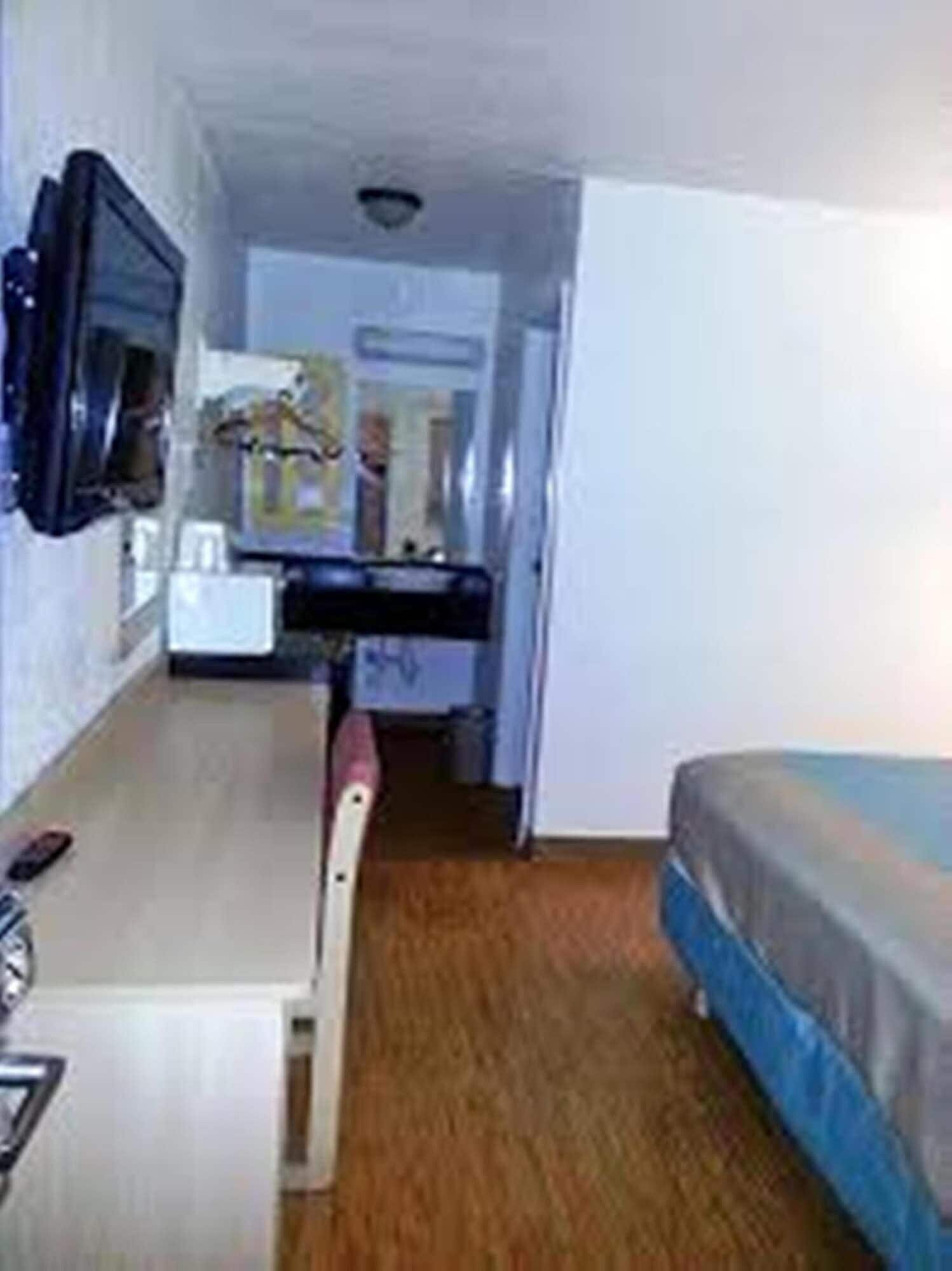 A small, compact room with a bed, a desk, and a chair. There is a television mounted on the wall and a window providing natural light.