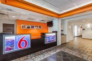 Un hall d’accueil d’hôtel avec un grand panneau portant le chiffre « 6 », un écran numérique et une pancarte indiquant : « Nous laisserons la lumière allumée pour vous. »