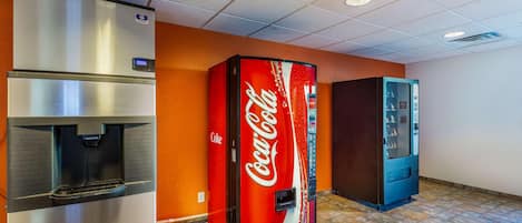 A vending machine, a water dispenser, and a refrigerator in a room with orange walls and a tiled floor.