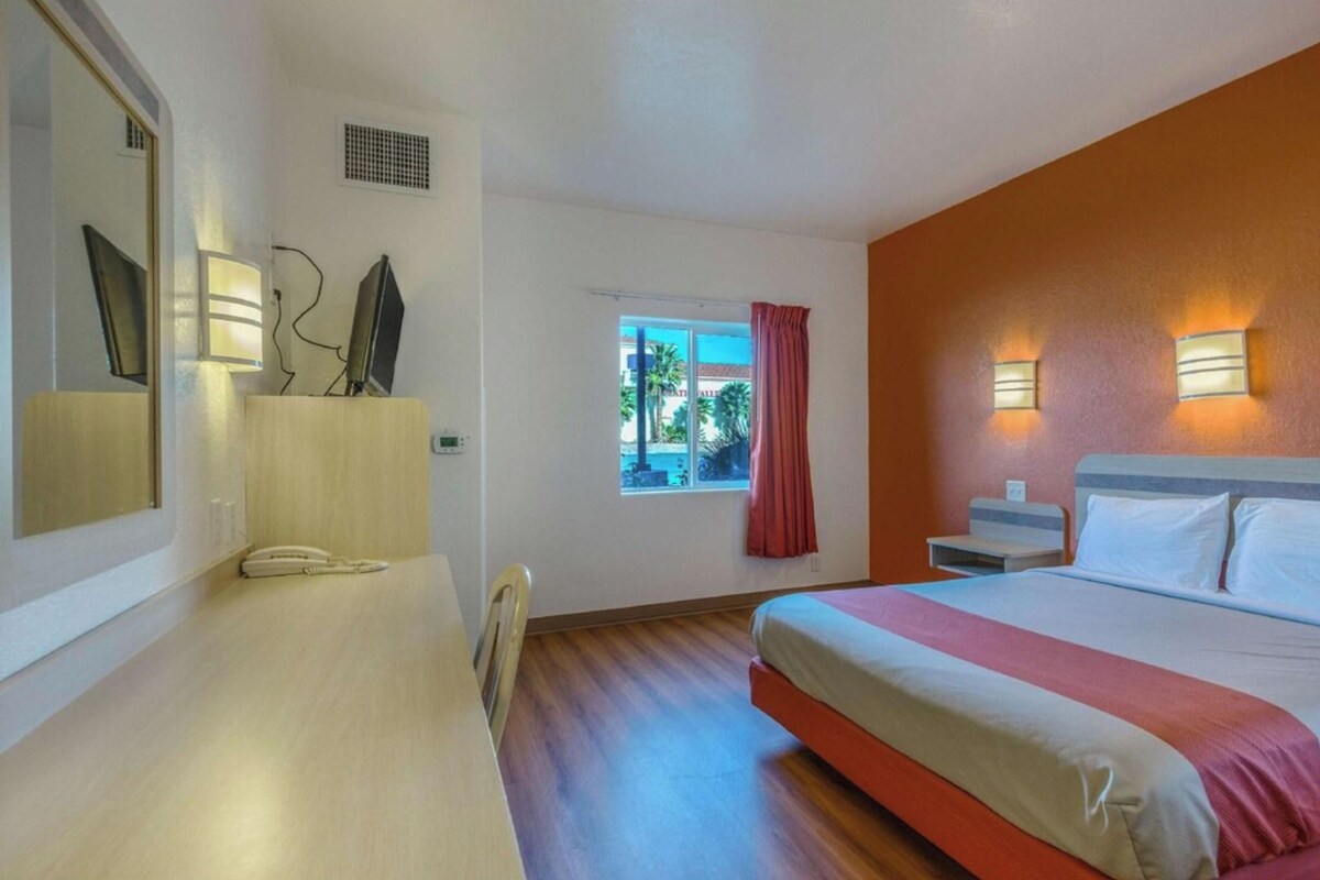 A hotel room with a bed, bedside table, lamp, window, and a view of the outside.