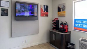 A modern office space with a mounted flat-screen TV displaying a news broadcast, a framed certificate, and a motivational poster. There are two coffee makers on a cabinet, and a trash bin is visible. - Motel 6 Lufkin, TX (Lufkin)