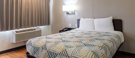 A hotel room with a bed, a bedside table, a lamp, an air conditioner, and a window with curtains.